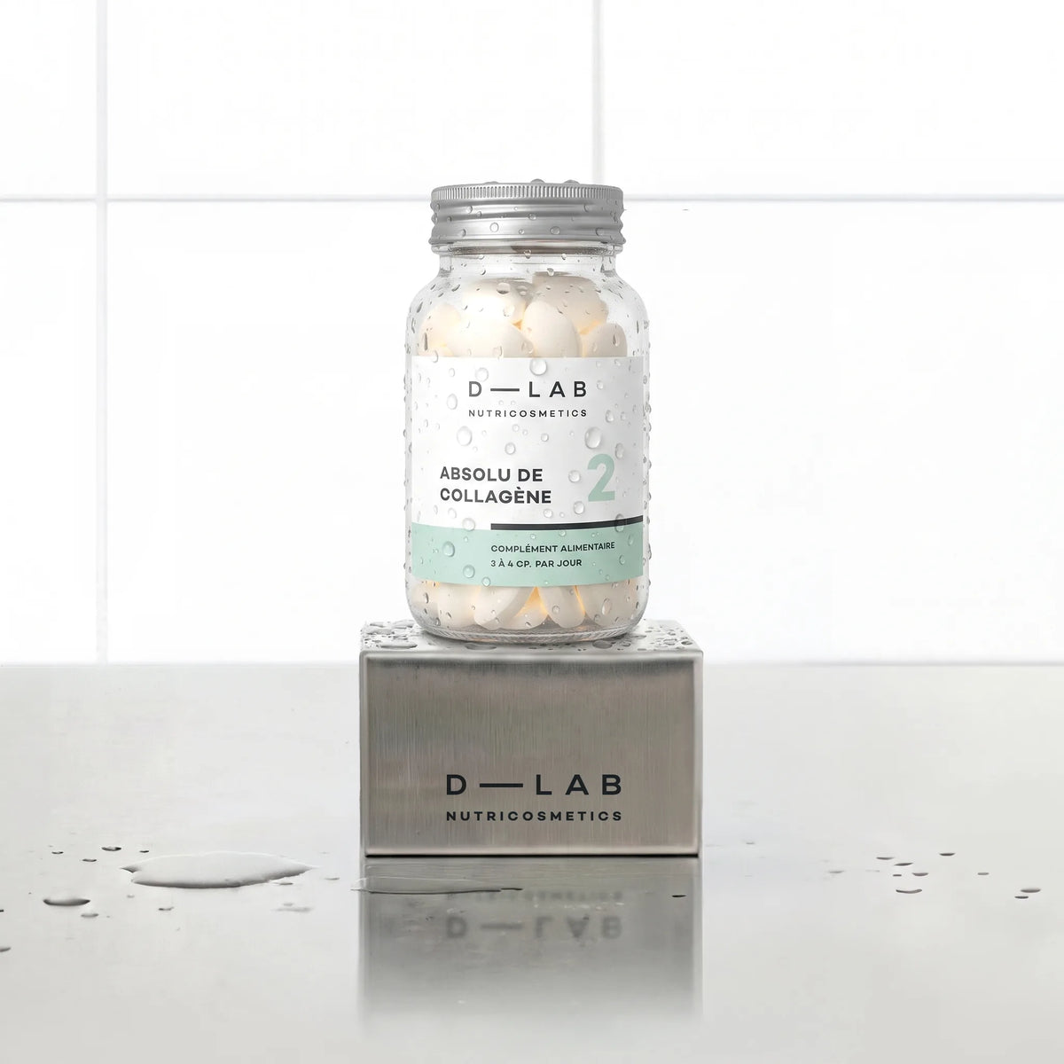 Absolu de Collagène D-LAB Nutricosmetics cure 1 mois, flacon en verre de comprimés perlé de gouttes d'eau posé sur un socle inox brossé, fond carrelage blanc