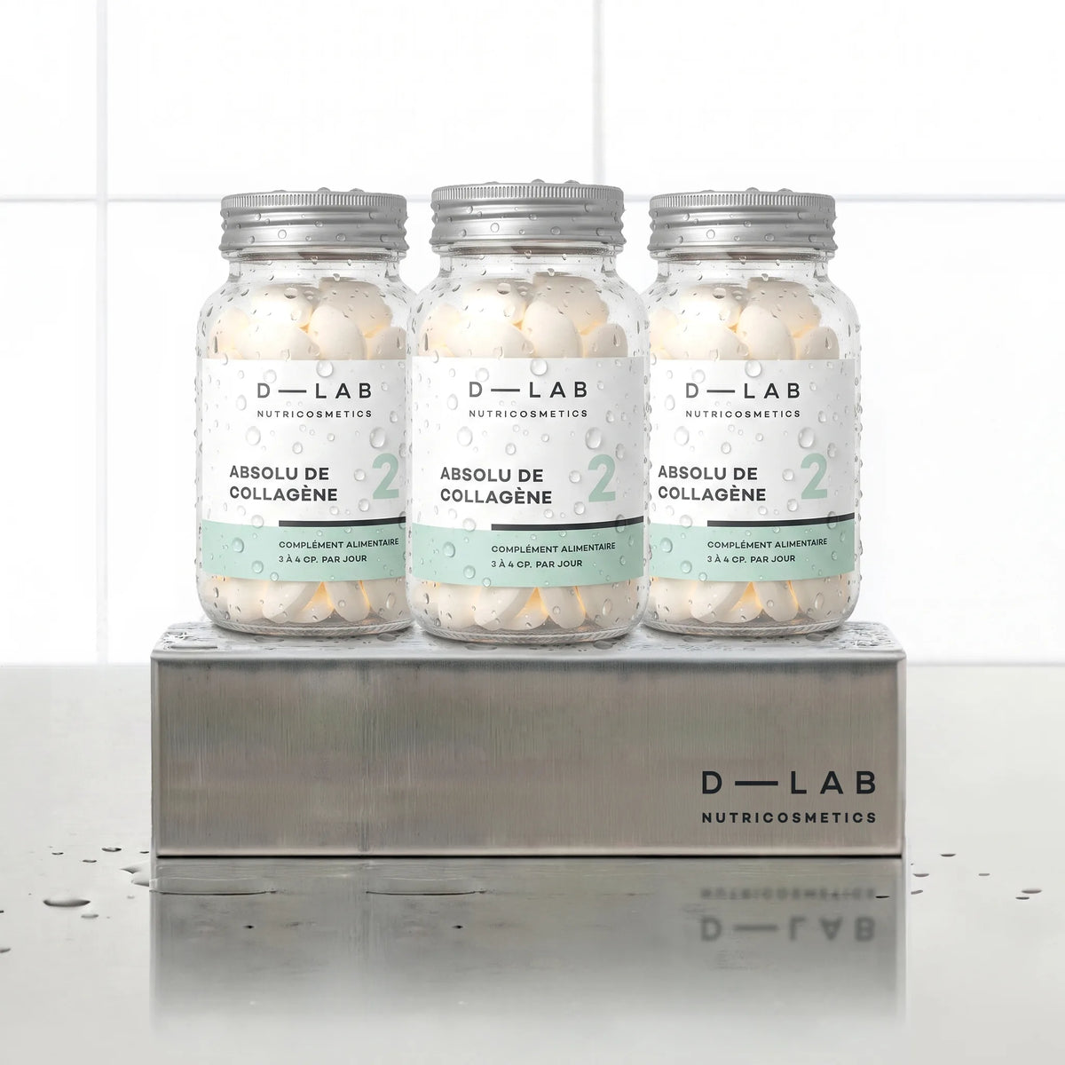 Absolu de Collagène D-LAB Nutricosmetics cure 3 mois, trois flacons en verre de comprimés alignés sur un socle inox brossé, décor carrelage blanc et reflets d'eau
