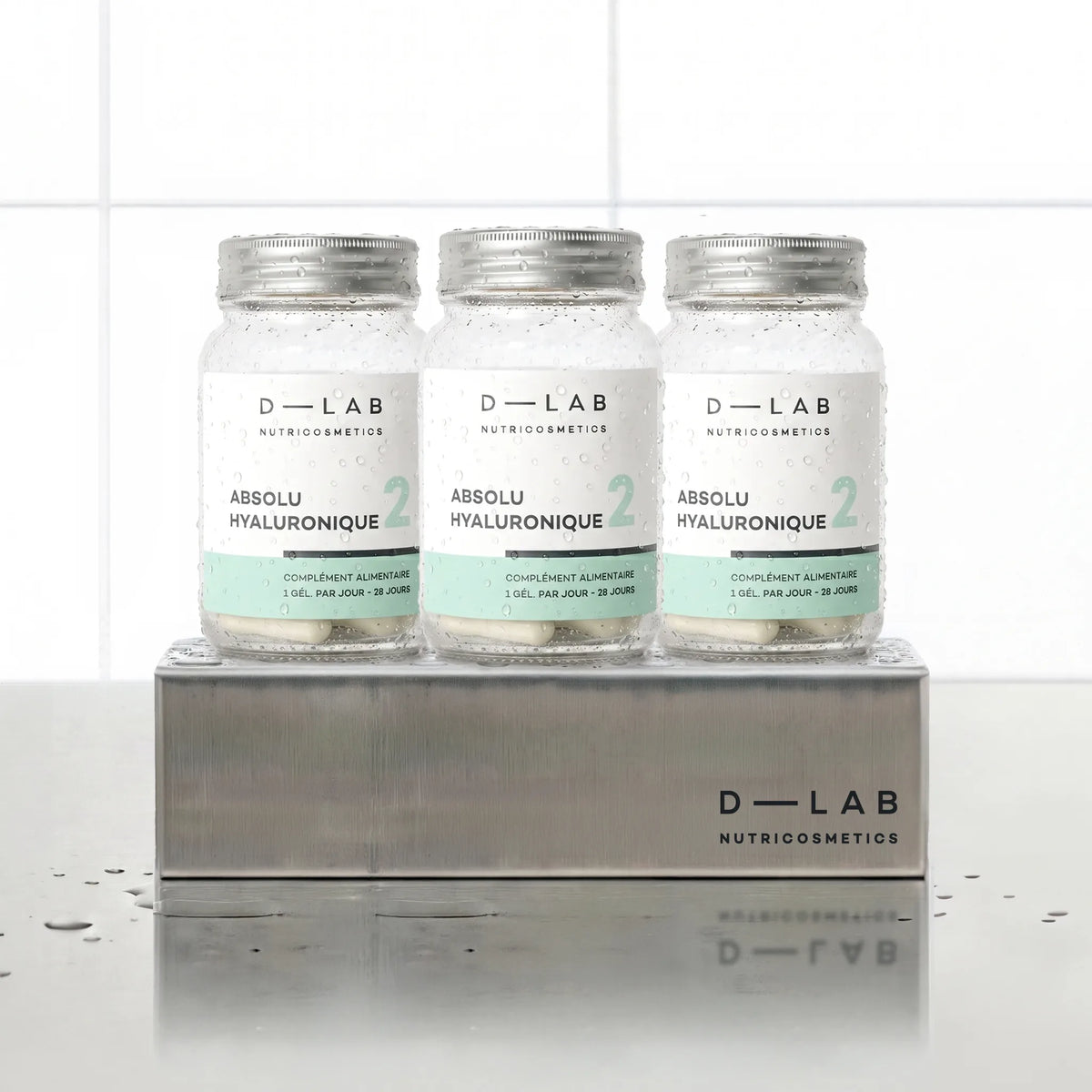Absolu Hyaluronique D-LAB Nutricosmetics, cure 3 mois : 3 flacons de gélules alignés sur un socle inox brossé, fond carrelage blanc