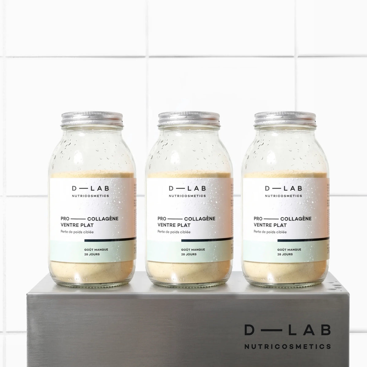Cure 3 mois Pro-Collagène Ventre Plat goût mangue D-LAB, trois flacons en verre alignés sur socle en métal brossé, fond carrelage blanc