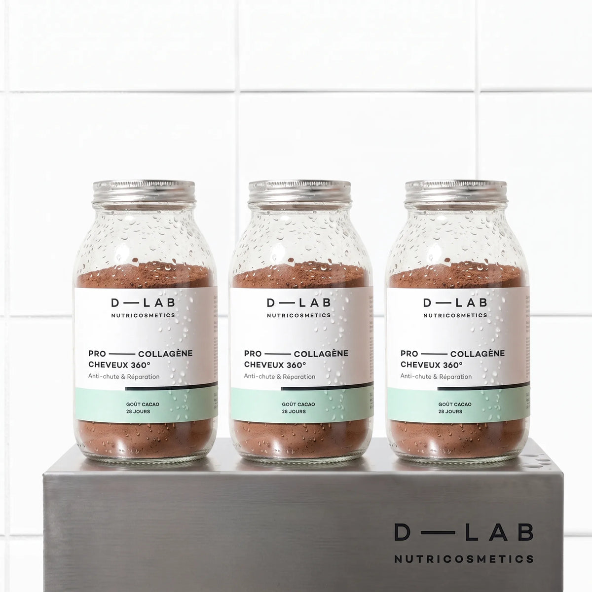 Cure 3 mois Pro-Collagène Cheveux 360 goût cacao D-LAB, trois flacons en verre alignés sur socle en métal brossé, fond carrelage blanc