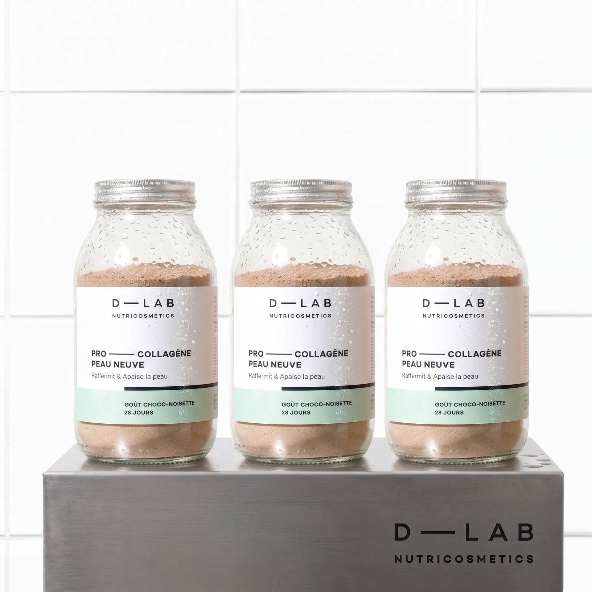 Cure 3 mois Pro-Collagène Peau Neuve goût choco-noisette D-LAB, trois flacons en verre alignés sur socle en métal brossé, fond carrelage blanc
