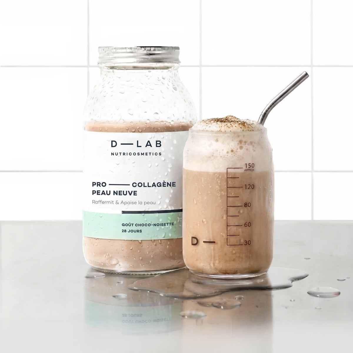 Flacon en verre Pro-Collagène Peau Neuve goût choco-noisette D-LAB et verre de boisson moussée saupoudrée de cacao, paille inox, plan de travail carrelé blanc