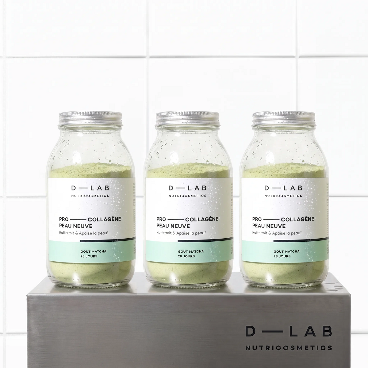 Cure 3 mois Pro-Collagène Peau Neuve goût matcha D-LAB, trois flacons en verre de poudre verte sur socle en métal brossé, fond carrelage blanc