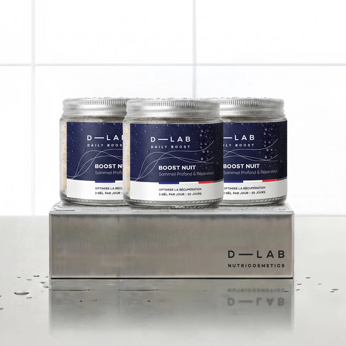 Boost Nuit Daily Boost D-LAB Nutricosmetics, cure 3 mois : 3 flacons de gélules alignés sur un socle inox brossé, fond carrelage blanc