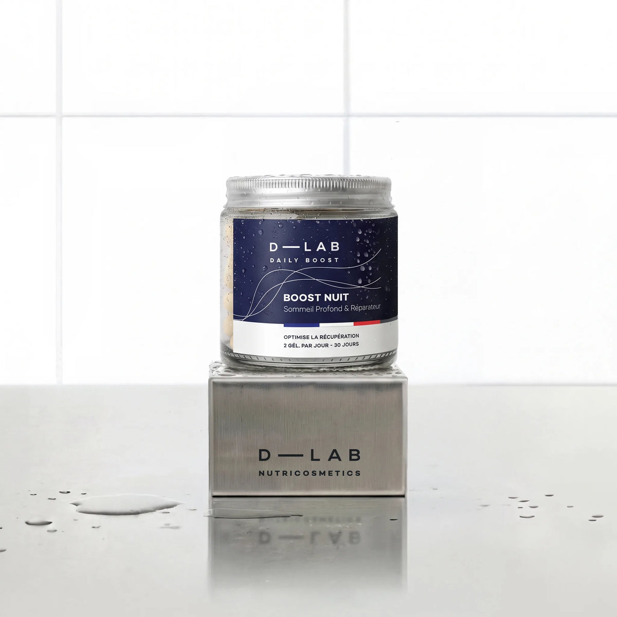 Boost Nuit Daily Boost D-LAB Nutricosmetics, cure 1 mois : flacon de gélules posé sur un socle inox brossé, fond carrelage blanc