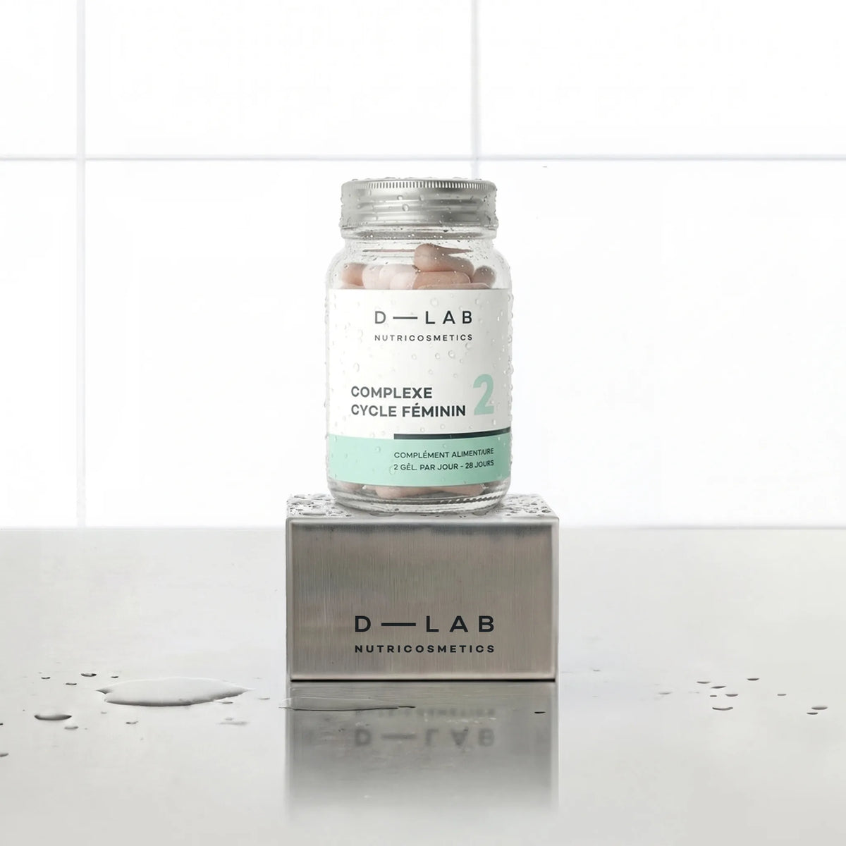Complexe Cycle Féminin D-LAB Nutricosmetics, cure 1 mois : flacon en verre de gélules posé sur un socle inox brossé, fond carrelage blanc