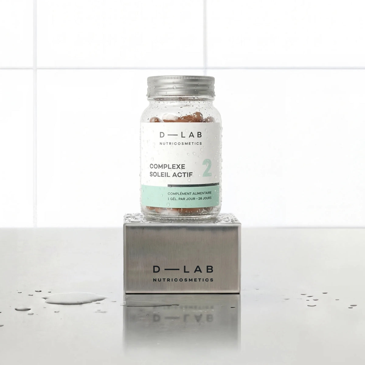 Complexe Soleil Actif D-LAB Nutricosmetics, cure 1 mois : flacon en verre de gélules posé sur un socle inox brossé, fond carrelage blanc