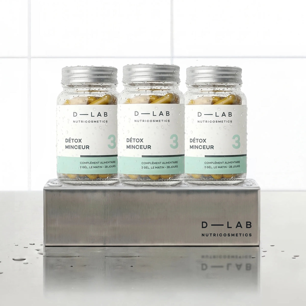 Détox Minceur D-LAB Nutricosmetics, cure 3 mois : 3 flacons de gélules alignés sur un socle inox brossé, fond carrelage blanc