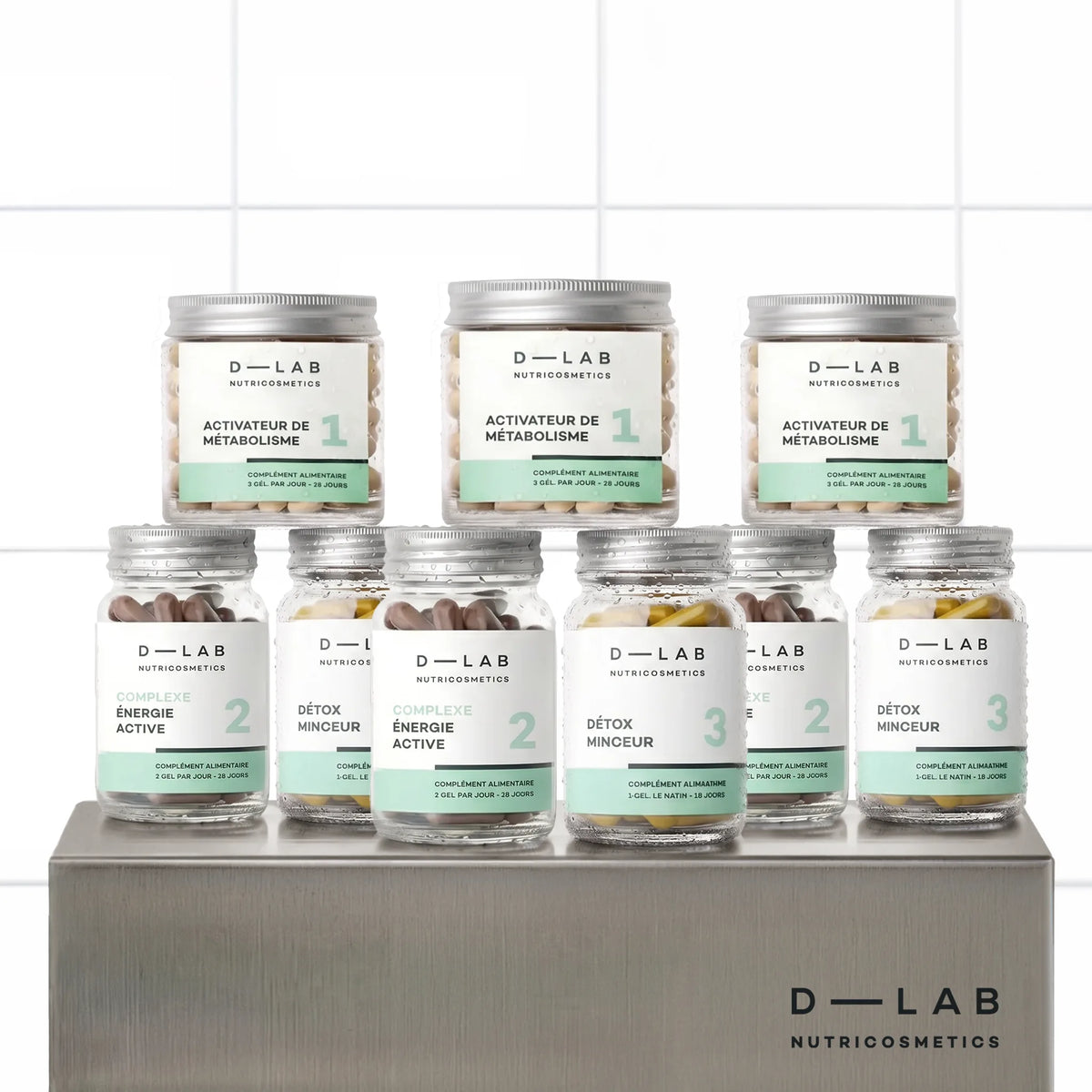 Routine Brûle-Graisses D-LAB cure 3 mois, neuf pots du programme en trois exemplaires alignés sur un socle inox brossé, fond carrelage blanc