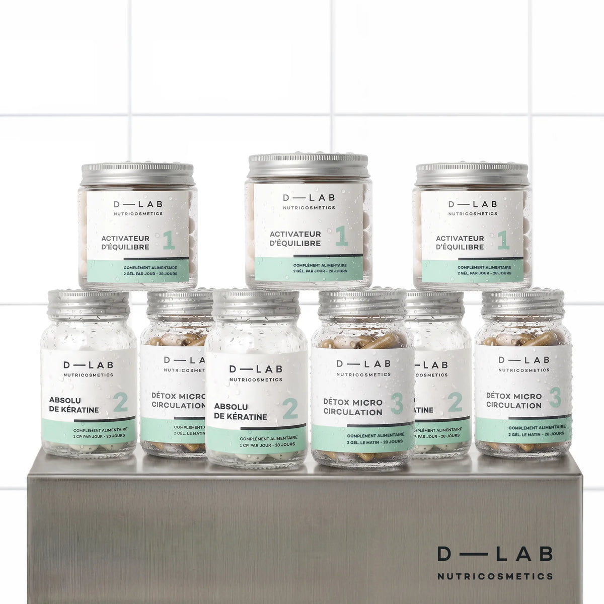 Routine Masse-Capillaire D-LAB cure 3 mois, neuf pots du programme en trois exemplaires alignés sur un socle inox brossé, fond carrelage blanc