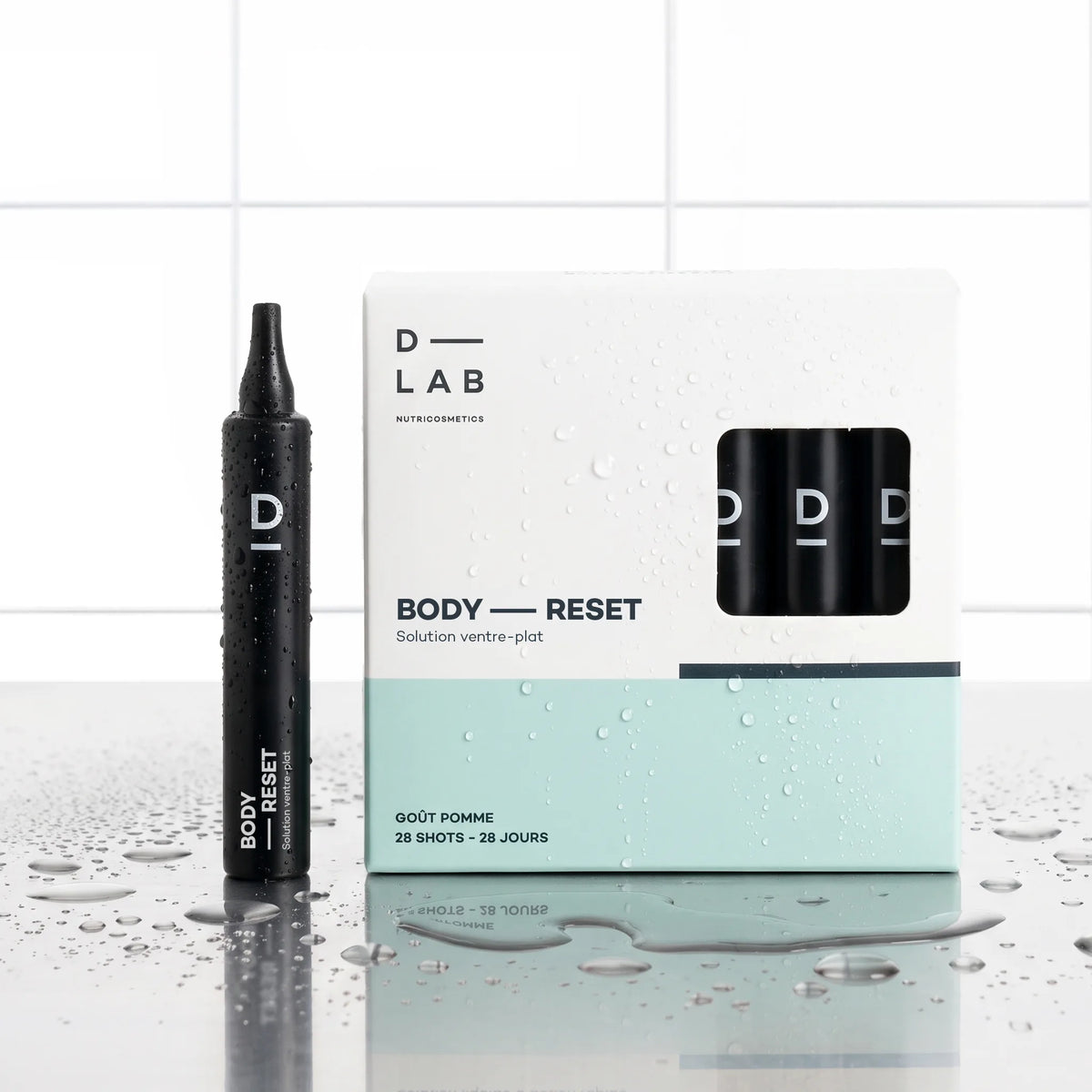 Solution Ventre-Plat Body-Reset D-LAB cure 28 jours, shot noir devant son étui 28 shots goût pomme, décor carrelage blanc et gouttes d'eau
