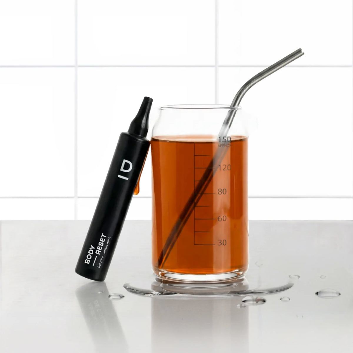 Solution Ventre-Plat Body-Reset D-LAB, shot noir liquide posé contre un verre gradué rempli de boisson goût pomme avec paille inox, fond carrelage blanc