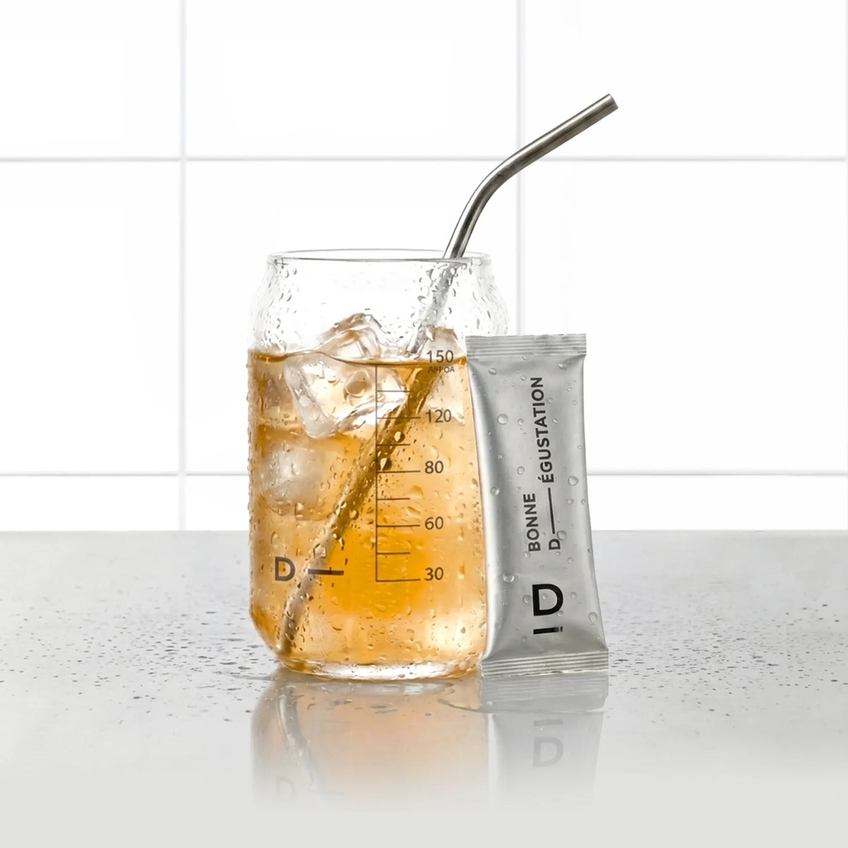 Sticks Ventre Plat Mangue D-LAB, stick de poudre posé contre un verre de boisson mangue glacée avec paille inox, fond carrelage blanc