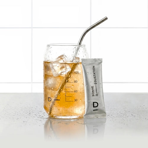 Sticks Ventre Plat Mangue D-LAB, stick de poudre posé contre un verre de boisson mangue glacée avec paille inox, fond carrelage blanc